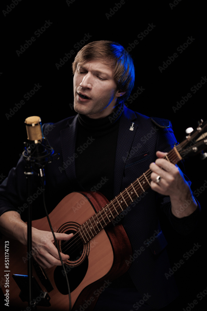 Fototapeta premium Male musician with guitar in hands playing and posing on black background in blue scenic light