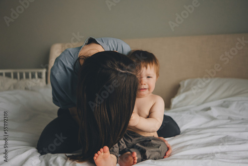 mom kisses a frustrated one-year-old baby sitting at home on the bed, mom's face is not visible behind her hair