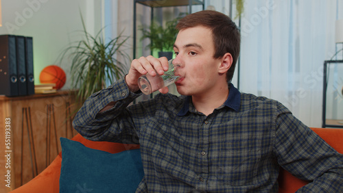 Portrait of thirsty teenager man sitting indoors holding glass of natural aqua make sips drinking still water preventing dehydration. Guy with good life habits, healthy slimming, weight loss concept