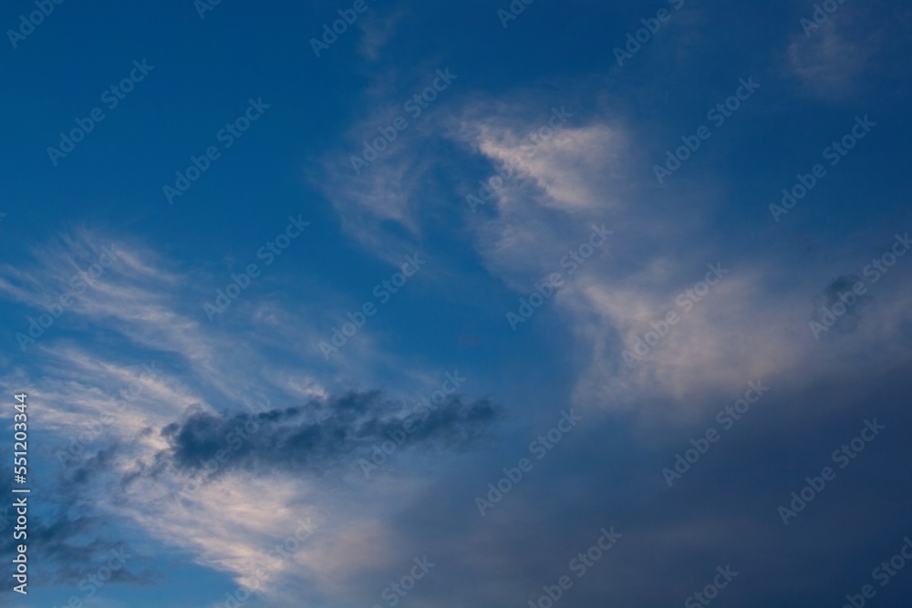 Dark wispy  clouds in front of thin white clouds in a blue sky