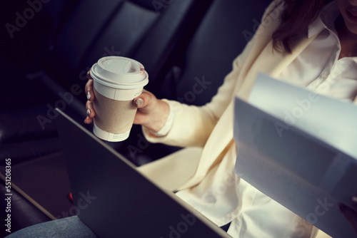 Close-up woman sitting in the back seat of the car with a paperwork in her hand and drinking coffee.
