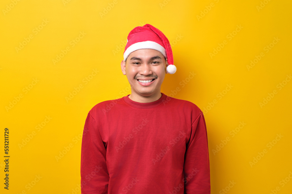 Friendly young Asian man in Santa hat smiling happily at camera on yellow studio background. celebration Christmas holiday and New Year concept