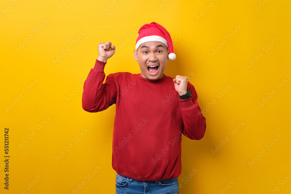 Excited young Asian man in Santa hat raising hands up, celebrating success on yellow studio background. celebration Christmas holiday and New Year concept Excited young Asian man in Santa hat raising hands up, celebrating success on yellow studio background. celebration Christmas holiday and New Year concept