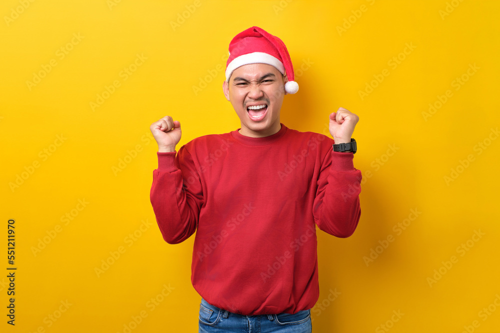 Naklejka premium Excited young Asian man in Santa hat raising hands up, celebrating success on yellow studio background. celebration Christmas holiday and New Year concept