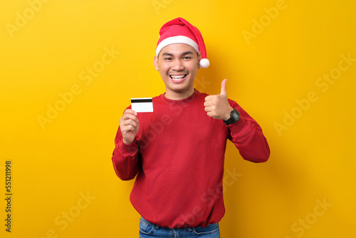 Cheerful young Asian man in Santa hat holding credit card and showing thumbs up on yellow studio background. celebration Christmas holiday and New Year concept © Sewupari Studio