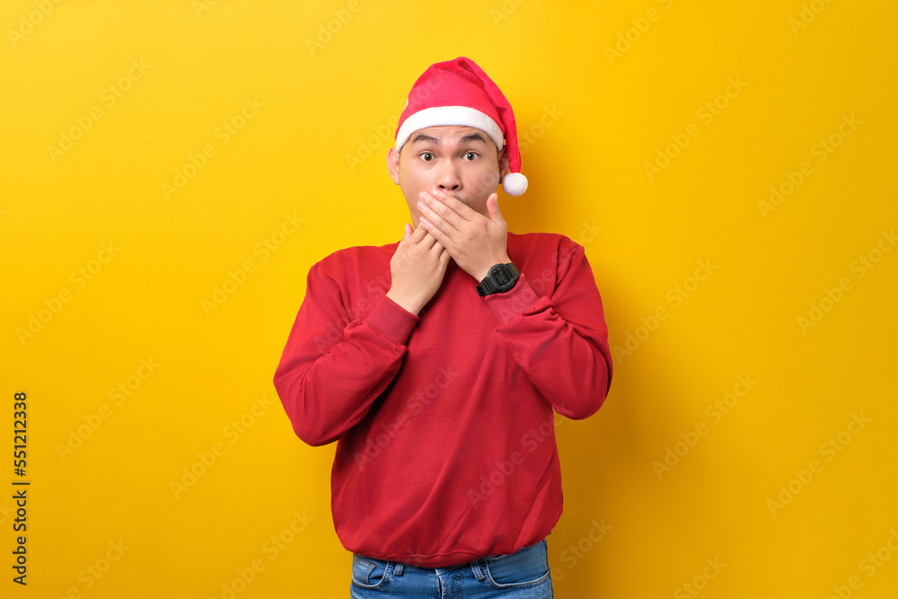Shocked young Asian man in Santa hat covering mouth with hands and looking at camera on yellow studio background. celebration Christmas holiday and New Year concept Shocked young Asian man in Santa hat covering mouth with hands and looking at camera on yellow studio background. celebration Christmas holiday and New Year concept
