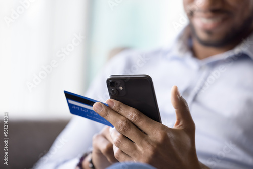 Focused mobile phone user man using credit card, ecommerce app on smartphone for buying, sitting on sofa at home, shopping on Internet, making online payment
