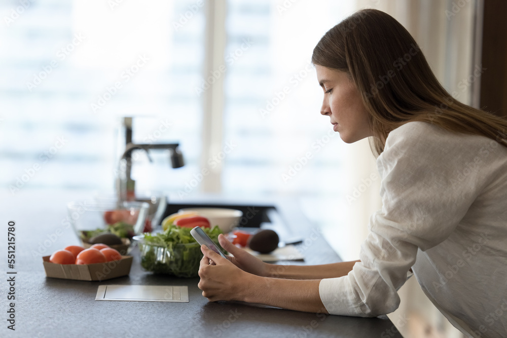 Serious focused chef girl using mobile phone at kitchen table, watching cook blogger video recipe, standing at table with fresh vegetables, natural food ingredients for salad, dinner