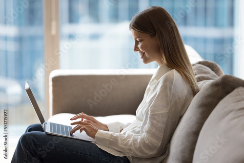 Happy cheerful pretty young freelance business woman working from home, using laptop computer, typing, chatting online, sitting on comfortable couch with big window in background. Side view