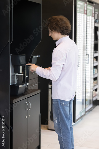 Vertical view, young man standing in modern office workspace during lunch break prepare morning hot coffee or tea beverage using professional vending machine indoor. Food and drink industry, lifestyle