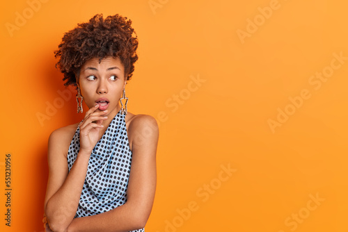 Studio shot of beautiful curly haired woman keeps mouth opened stares impressed at something awesome reacts to shocking news wears fashionable clothes isolated over orange background copy space