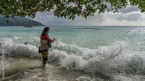 A man stands in the turquoise ocean near the shore. The surf waves are foaming. Splashes in the air. Green branches hang over the beach. A hill in the distance. Clouds in the sky. Seychelles. Mahe.