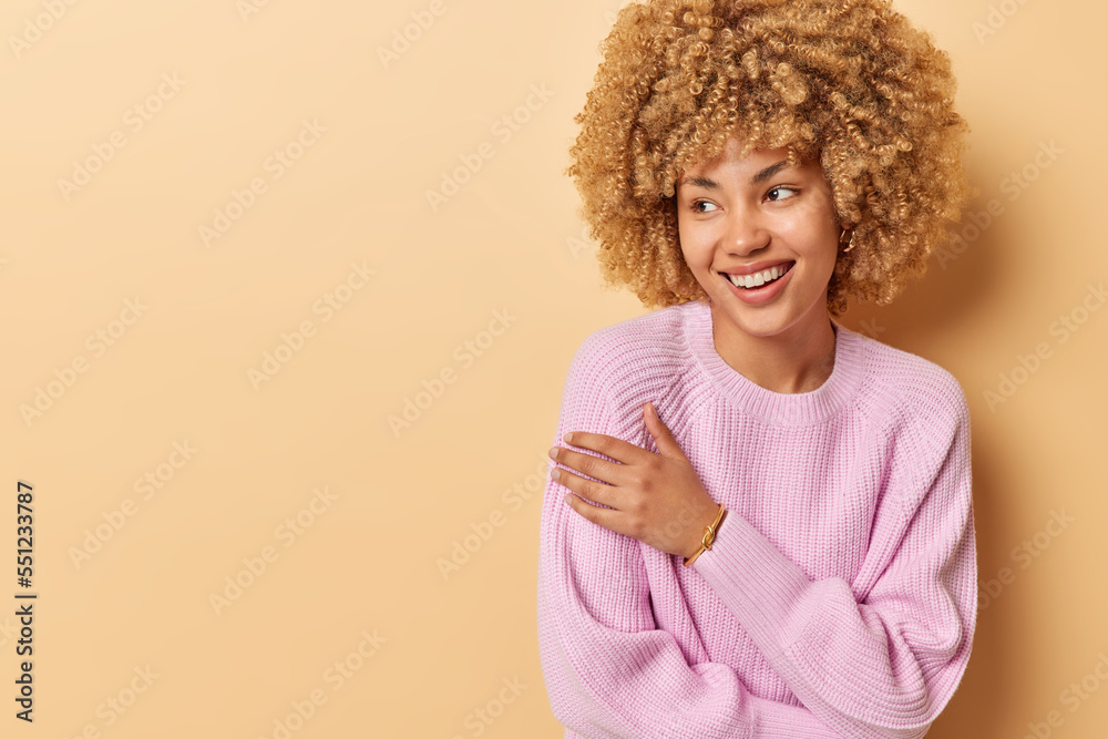 Waist up shot of pretty curly haired woman keeps arms crossed smiles gently looks aside recalls something nice from her life isolated over beige background blank space for your promotional text Waist up shot of pretty curly haired woman keeps arms crossed smiles gently looks aside recalls something nice from her life isolated over beige background blank space for your promotional text