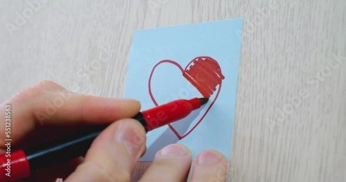 A man's hand draws a red heart on a blue sticky note with a marker. Concept of love, creativity, psychology, and relationships