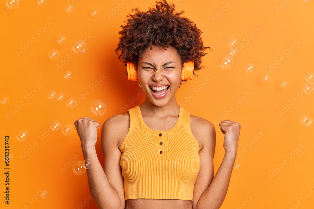 Indoor shot of positive woman with dark curly hair clenches fists celebrates success listens music via headphones dressed in casual cropped top isolated over orange background. Yes I did it. Indoor shot of positive woman with dark curly hair clenches fists celebrates success listens music via headphones dressed in casual cropped top isolated over orange background. Yes I did it.