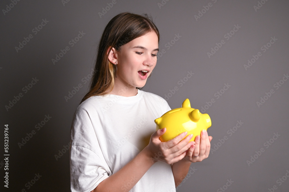 Surprised teenage girl holding piggy bank. Young girl with yellow piggy bank on gray background Surprised teenage girl holding piggy bank. Young girl with yellow piggy bank on gray background