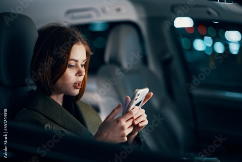 horizontal portrait of a stylish, luxurious woman in a leather coat sitting in a black car at night in the passenger seat, emotionally looking at her smartphone during the trip with her mouth open © SHOTPRIME STUDIO