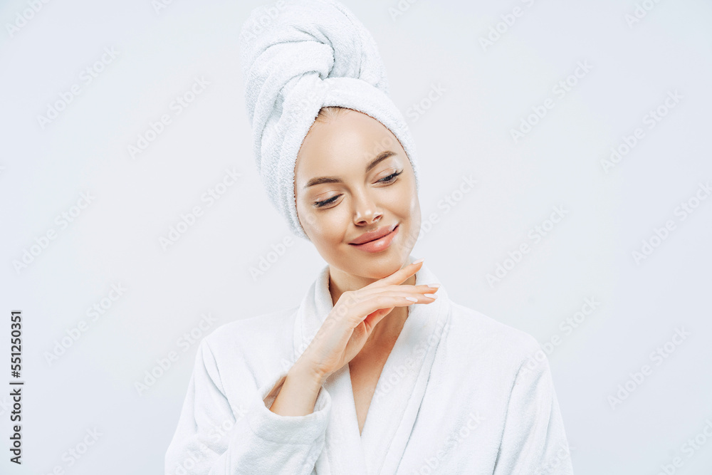 Pretty natural woman touches gently jawline, wears bath towel wrapped on head, dries hair after shower, cares about body, has clean skin, prepares for evening party, dressed in domestic robe Pretty natural woman touches gently jawline, wears bath towel wrapped on head, dries hair after shower, cares about body, has clean skin, prepares for evening party, dressed in domestic robe