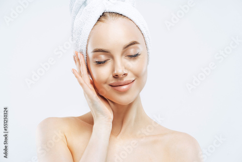 Pretty calm woman with wrap towel on head, closes eyes, enjoys softness of skin, undergoes skin care treatment, stands bare shoulders indoor, relaxed after bath, isolated over white background