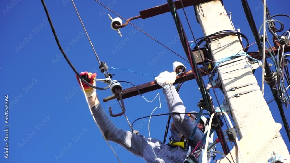 Skilled electrician in helmet fixes wires standing on ladder near high pole against blue sky on summer day backside view. Electrical service and mounting on the pole. Slow motion Skilled electrician in helmet fixes wires standing on ladder near high pole against blue sky on summer day backside view. Electrical service and mounting on the pole. Slow motion