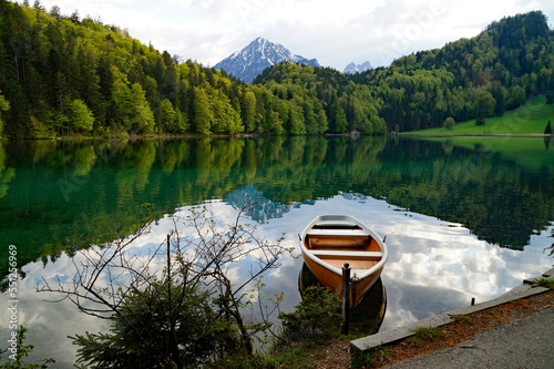 a boat resting on the emerald-green waters of lake Alatsee in Fuessen with the snowy Bavarian Alps reflected in the calm water and the lush green spring forest in the background, Bavaria, Germany photo