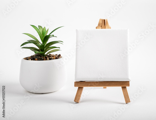 Blank white canvas mock up, empty paper on mini wood tripod, easel and potted house plant on desk