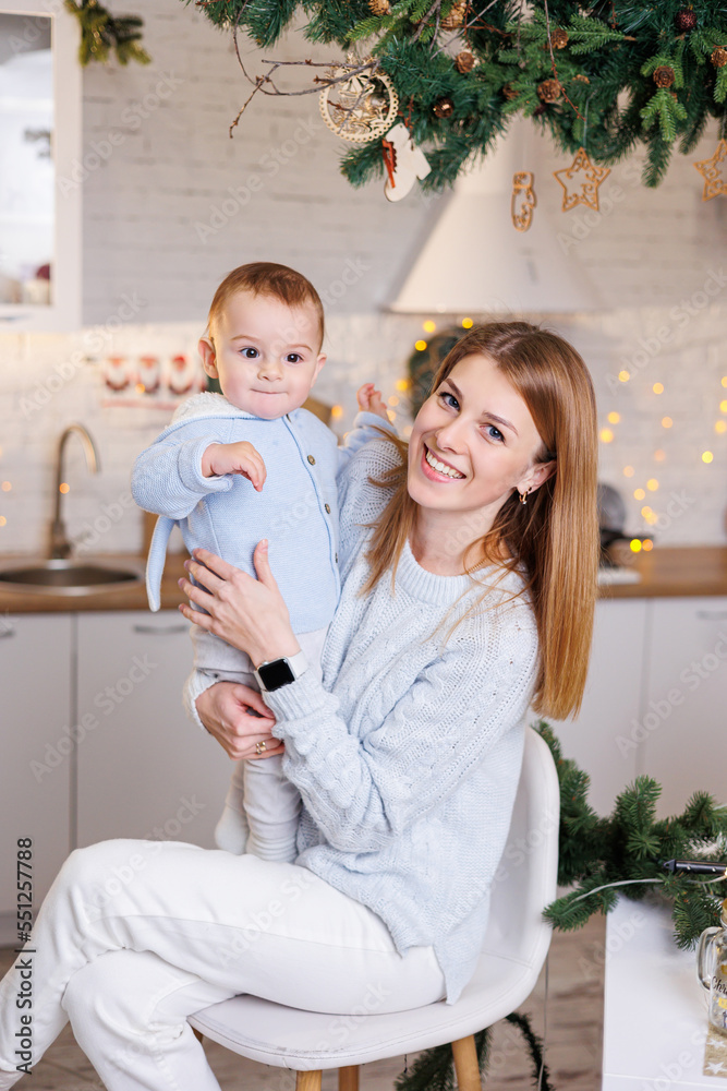 A young happy mother with her son in her arms welcomes the new year. A woman with a child on the background of a New Year's kitchen at home A young happy mother with her son in her arms welcomes the new year. A woman with a child on the background of a New Year's kitchen at home
