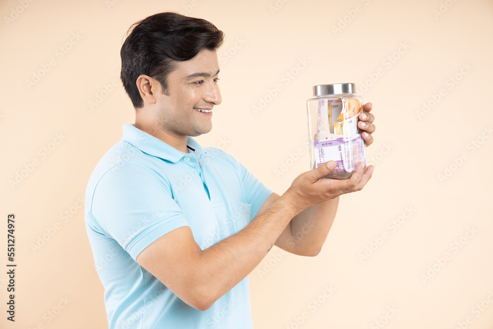 Portrait of happy Indian man holding glass jar full of rupee notes isolated on beige studio background. Money saving and investing concept. Portrait of happy Indian man holding glass jar full of rupee notes isolated on beige studio background. Money saving and investing concept.
