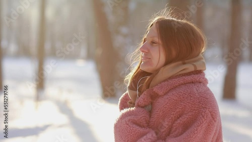 Dreamy young woman with frosty breath in winter park, listening to music is using headphones. Girl in winter fluffy pink coat and knitted mittens having fun outdoors in cold snowy day with sunlight