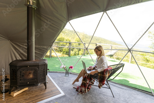 Young caucasian woman using and watching on laptop computer on in dome camping hotel. Tourism, vacation and weekend.