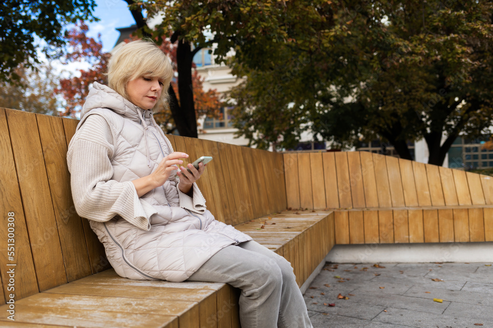 Free Wi-Fi in city park. Adult Caucasian woman is sitting on bench and using smartphone. Copy space. Concept of online communication and social web Free Wi-Fi in city park. Adult Caucasian woman is sitting on bench and using smartphone. Copy space. Concept of online communication and social web