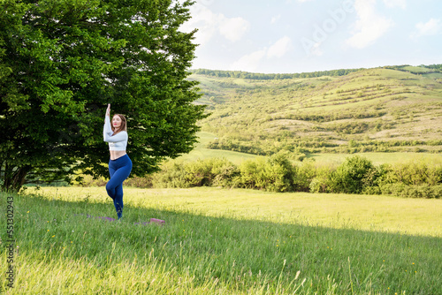 Young woman in sporty outfit exercising yoga and stretching on mat in meadow outdoors. Hills landscape on background. Healthy active lifestyle in summer