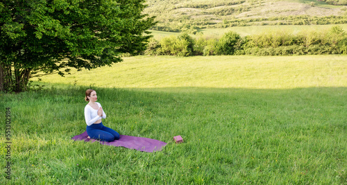 Young woman in sporty outfit relaxing and meditating on mat in sunset meadow outdoors. Hills landscape on background. Healthy active lifestyle in summer