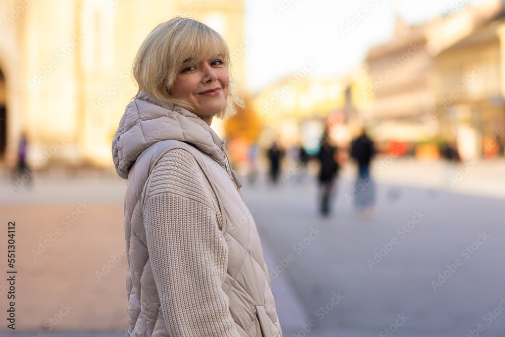 Portrait of Caucasian adult smiling blonde woman in jacket posing in city centre. Concept of carefree and psychology