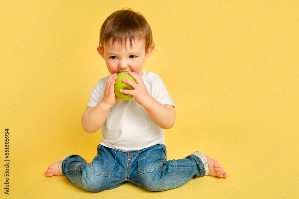 Happy toddler baby eating a green apple on studio yellow background in full length. Child with a big green apple in a white t-shirt and blue jeans, copy space. Kid aged one year and four months Happy toddler baby eating a green apple on studio yellow background in full length. Child with a big green apple in a white t-shirt and blue jeans, copy space. Kid aged one year and four months