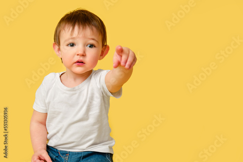 Portrait of upset toddler baby on studio yellow background. Offended child points forward with index finger in white t-shirt and blue jeans, copy space. Kid aged one year and four months