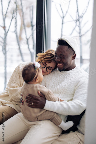 Happy interracial family sits on windowsill and plays with their baby daughter against background of window. Concept of interracial family and unity between different human races. baby daughter. © Stanislav