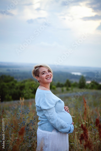 young pregnant woman smiles and hugs her stomach against the backdrop of nature