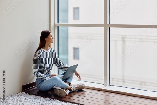 A woman freelancer sits with her laptop full-length at a large window, looking out at tall buildings in the city, work, training and shopping online © SHOTPRIME STUDIO