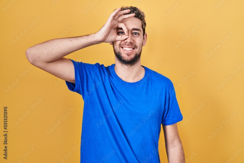Hispanic man with beard standing over yellow background doing ok gesture with hand smiling, eye looking through fingers with happy face. Hispanic man with beard standing over yellow background doing ok gesture with hand smiling, eye looking through fingers with happy face.