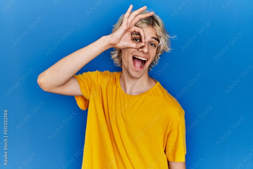 Young modern man standing over blue background doing ok gesture with hand smiling, eye looking through fingers with happy face.