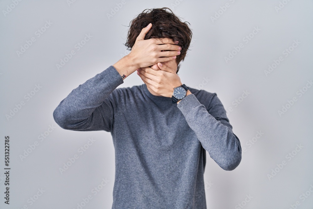 Young man standing over isolated background covering eyes and mouth with hands, surprised and shocked. hiding emotion Young man standing over isolated background covering eyes and mouth with hands, surprised and shocked. hiding emotion