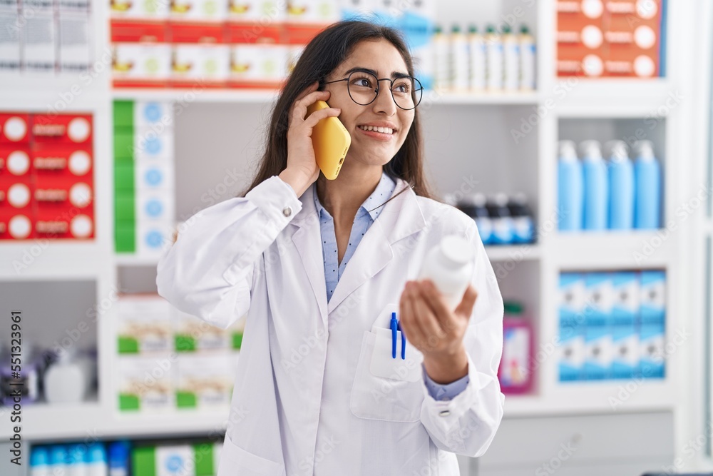 Young hispanic girl pharmacist talking on the smartphone holding pills at pharmacy