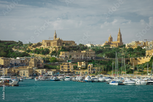 Entrance to the Mgarr port on the island of Gozo with visible marina, churches and boats moored. Beautiful cityscape of seaside town.
