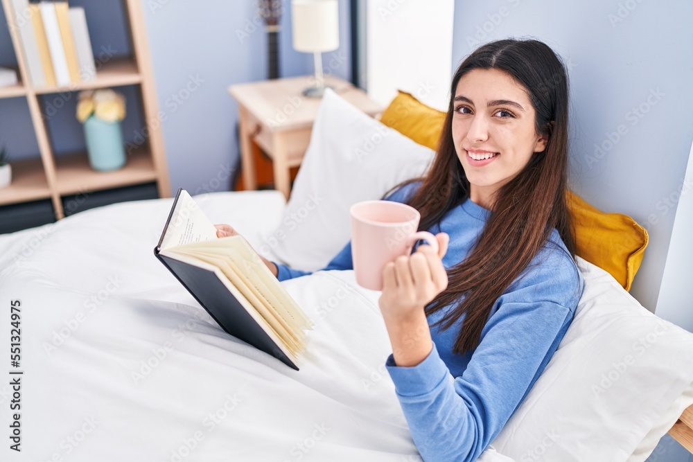 Young beautiful hispanic woman drinking cup of coffee reading book at bedroom