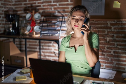 Young beautiful woman working at the office at night speaking on the phone relaxed with serious expression on face. simple and natural looking at the camera.