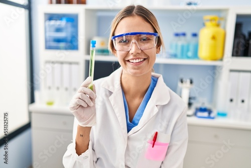 Young blonde woman working at scientist laboratory holding sample looking positive and happy standing and smiling with a confident smile showing teeth
