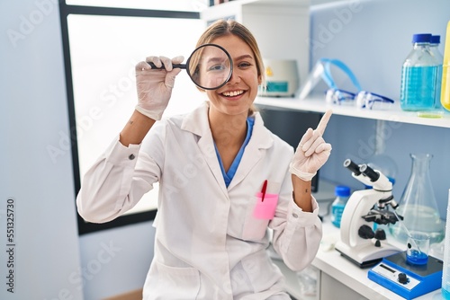 Young blonde woman working at scientist laboratory using magnifying glass smiling happy pointing with hand and finger to the side