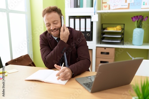 Middle age man business worker talking on smartphone writing on notebook at office