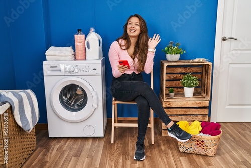 Young hispanic woman sitting waiting for laundry using smartphone waiving saying hello happy and smiling, friendly welcome gesture